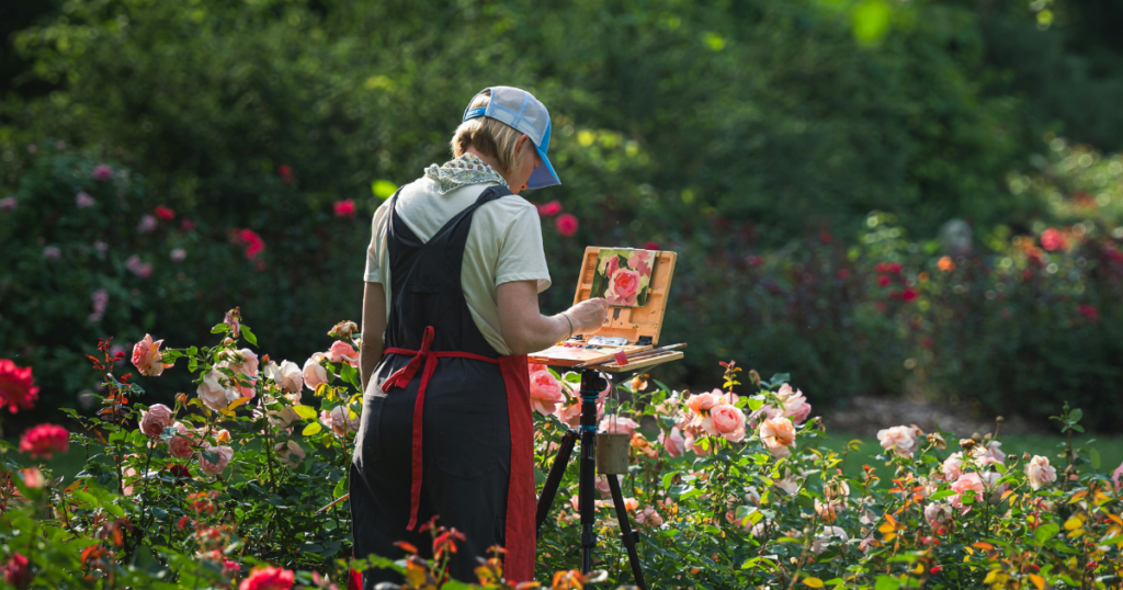 Easy Canvas Art Ideas An artist wearing a baseball cap, overalls, and a red apron stands in a lush garden filled with blooming pink and red roses. They are using a portable pochade box easel mounted on a tripod to paint a small floral study of the roses in front of them.