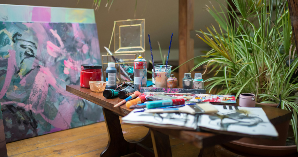 Easy Canvas Art Ideas A close-up of a wooden workbench cluttered with art supplies, including tubes of acrylic paint, brushes, spray cans, and an open sketchbook.